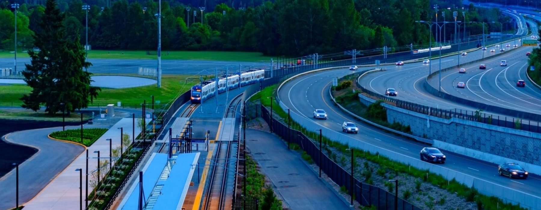 A light rail station and approaching train run alongside a curved multi-lane highway at dusk, with dense evergreen forest and a colorful sunset sky in the background.